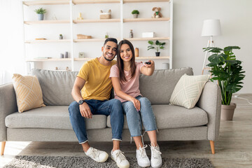 Obraz premium A young couple sits comfortably on a soft couch in a modern living area, smiling and holding a remote control. The warm atmosphere is enhanced by plants and decorative shelves.
