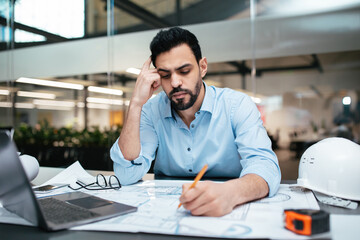 Naklejka na ściany i meble A man in a light blue shirt is seated at an office desk, deeply engaged in sketching plans. He is surrounded by engineering tools and a laptop, indicating intense focus on his work. Naklejka na ściany i meble A man in a light blue shirt is seated at an office desk, deeply engaged in sketching plans. He is surrounded by engineering tools and a laptop, indicating intense focus on his work.