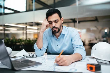 Fotobehang Muziek A man in a light blue shirt is seated at an office desk, deeply engaged in sketching plans. He is surrounded by engineering tools and a laptop, indicating intense focus on his work.  © Prostock-studio