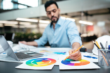 A designer focuses on assembling color wheels using pencils and papers in a modern office environment. Tools and plans are spread across the table, showcasing creativity.
