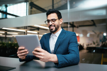 A man in a blazer sits at a sleek desk in a contemporary office. He smiles as he interacts with his tablet, surrounded by glass walls and plants, showing a productive environment.