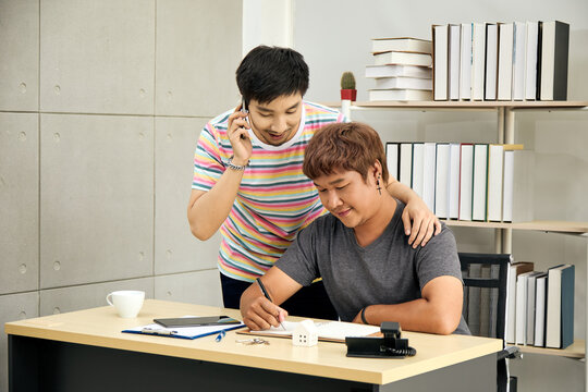 Asian gay couple working together at desk in home office, one man on phone while other takes notes. Concept of LGBTQ+ teamwork, support, communication, modern lifestyle, and remote business.