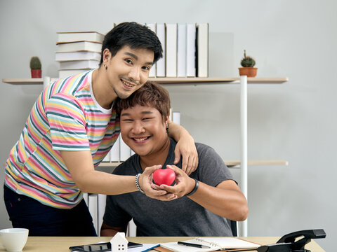 Asian gay couple smiling and holding a red heart together while sitting at home office desk. Concept of LGBTQ+ love, relationship support, health awareness, care, and emotional connection