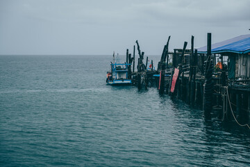 Small fishing boat sailing away from a weathered pier, gliding across the calm sea under a blanket of clouds on a tranquil day on Koh Kood, Thailand