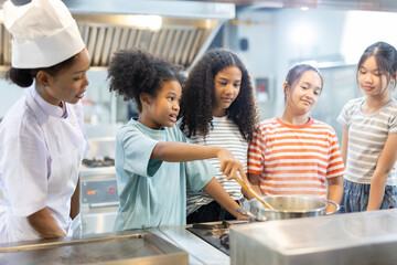 Professional female chef teaching diverse group of kids how to cook in modern kitchen. Concept of culinary education, teamwork, multicultural learning and healthy cooking skills for children.
