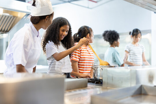 Professional female chef teaching diverse group of kids how to cook in modern kitchen. Concept of culinary education, teamwork, multicultural learning and healthy cooking skills for children.