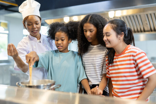 Professional female chef teaching diverse group of kids how to cook in modern kitchen. Concept of culinary education, teamwork, multicultural learning and healthy cooking skills for children. - Powered by Adobe