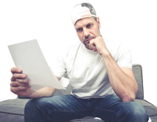 Thoughtful man in cap sits on gray couch, studying a blank white paper, hand on chin, looking at the document