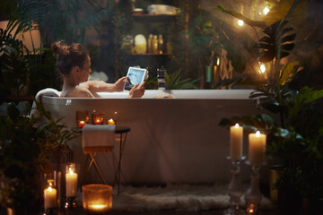 Woman in bubble bath holding a tablet, surrounded by candles and greenery. Evokes relaxation, luxury, and modern self-care with a tech twist.