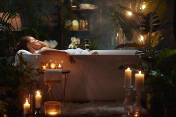A woman with her hair in a bun relaxes in a bubble bath, amidst plants and candles, in a softly lit bathroom. The scene evokes relaxation and luxurious self-care.
