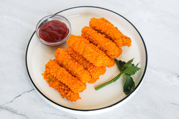Several crispy, golden-orange chicken nugget sticks, perfectly arranged on a white plate alongside a small glass bowl of vibrant red ketchup. Captured with natural light on a marble background.