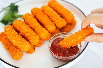 Hand dipping a crispy, golden-orange chicken nugget stick into a red ketchup, with a blurred background of more nuggets on a white plate. Captured with natural light on a marble background.