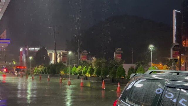 Rainy night scene on an urban street with wet road reflections, traffic cones, parked car and glowing city lights.