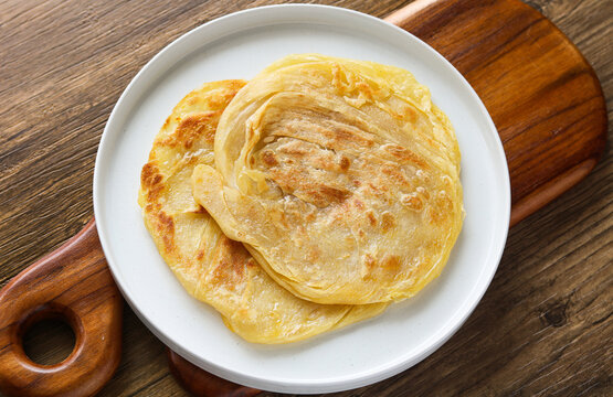 Two pieces of golden-brown Roti Canai (or Paratha) served on a white plate. Captured with natural light on a wooden background.