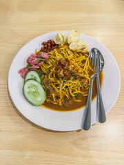 A plate of traditional Indonesian Mie Aceh served with rich spicy curry sauce, beef, cucumber slices, peanuts, crackers, and fried shallots on top.