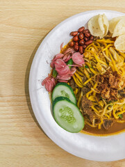 A plate of traditional Indonesian Mie Aceh served with rich spicy curry sauce, beef, cucumber slices, peanuts, crackers, and fried shallots on top.