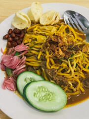 A plate of traditional Indonesian Mie Aceh served with rich spicy curry sauce, beef, cucumber slices, peanuts, crackers, and fried shallots on top.