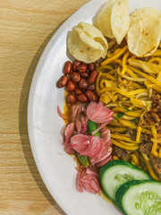 A plate of traditional Indonesian Mie Aceh served with rich spicy curry sauce, beef, cucumber slices, peanuts, crackers, and fried shallots on top.