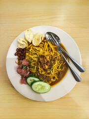 A plate of traditional Indonesian Mie Aceh served with rich spicy curry sauce, beef, cucumber slices, peanuts, crackers, and fried shallots on top.