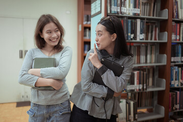 Two young women smiling and chatting happily in a library while holding books and tablets