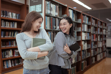 Two young women smiling and chatting happily in a library while holding books and tablets