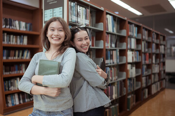 Two young women smiling and chatting happily in a library while holding books and tablets