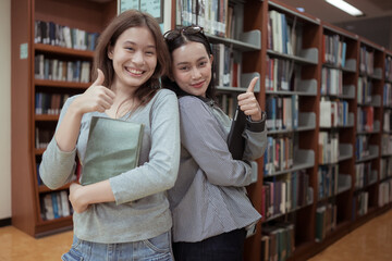 Two young women smiling and chatting happily in a library while holding books and tablets