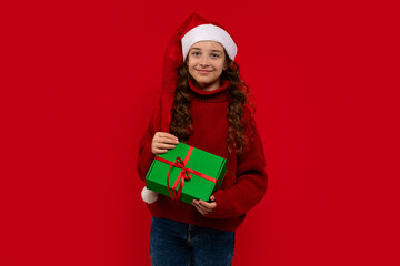 Smiling girl in a Santa hat and red sweater holds a green Christmas gift with a red ribbon