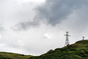 Powerline towers on green hillside beneath stormy sky. Electric transmission towers rise above lush vegetation while storm clouds loom, contrasting nature and human-made infrastructure