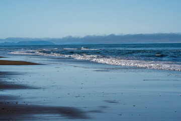 Blue ocean waves rolling calmly toward sandy shoreline. A calm ocean stretches to the horizon as waves gently roll onto the sandy shoreline under a bright blue sky with distant mist clouds