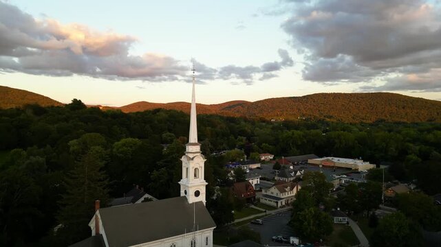 Aerial rising shot of White First Congregational Church in American town with golden green mountains at sunset time. Lee Town, Massachusetts, United States in Autumn Season. Panorama wide shot.