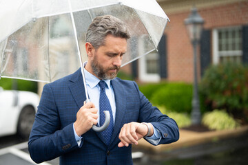 Businessman waits under the rain holding an umbrella. A confident man checks his watch while standing on a wet street. Businessman checks the time. Man watching the clock. A punctual business man.