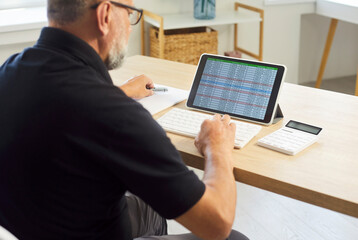 Senior man accountant reviews spreadsheet on tablet. At a desk, he analyzes finance and business accounting with a calculator at home office. Remote money management and budgeting concept.