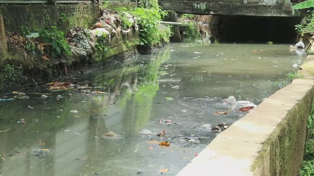 A neglected urban canal scene shows dirty, polluted water littered with plastic and debris, flanked by concrete walls and a fence. Greenery along the banks adds contrast to the grim setting.