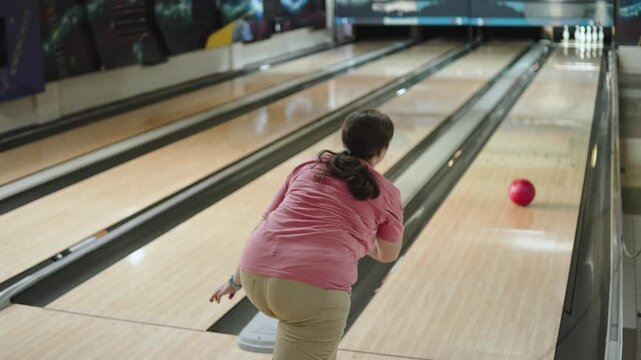 Caucasian woman at bowling alley throws ball and knocks down couple of pins. Bowler throw is not very successful, and bowling ball rolls down one of lanes.