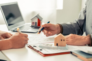 Signing a contract next to a model home, symbolizing home ownership. A real estate agent shows a small model home to a client with a calculator. Contract documents on a table.