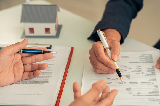Signing a contract next to a model home, symbolizing home ownership. A real estate agent shows a small model home to a client with a calculator. Contract documents on a table.