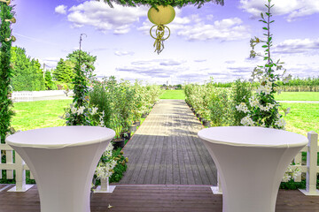 Zwei elegante Stehtische heißen die Gäste willkommen, bevor ein herrlicher Holzweg, gesäumt von frischem Grün und weißen Blüten, in die weite Natur führt. Oben glänzt eine goldene Dekoration – ein Sym