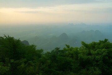Misty mountain landscape at sunrise with soft golden light illuminating the hazy sky above lush green forest canopy and distant valley