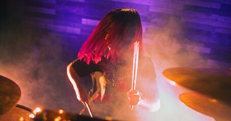 Professional female drummer with red hair performs on the stage of nightclub. Girl plays drums masterfully with drumsticks. The performance is accompanied by multi-colored light show