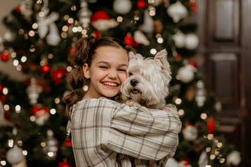 Close-up portrait of a joyful young girl with pigtails, hugging her small white Westie dog and smiling brightly in front of a sparkling, decorated Christmas tree, capturing holiday cheer