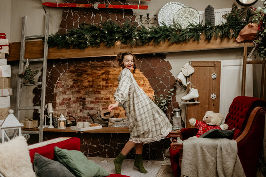 Joyful young girl in a cozy plaid dress and knit socks twirling happily in a rustic living room with a brick fireplace, beautifully decorated for Christmas and the holiday season