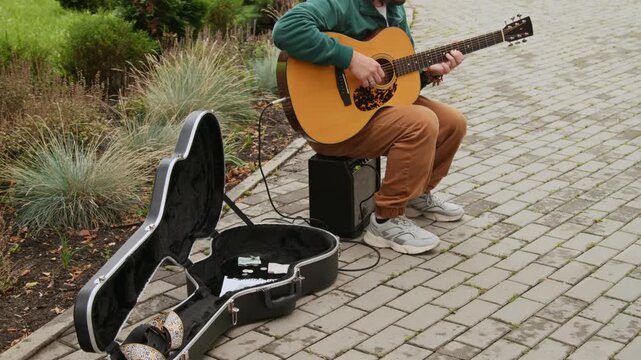 Medium full shot of male street musician sitting on sidewalk in green urban park, playing solo on guitar, next to instrument case with coins and banknotes, while collecting donations from public