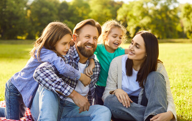 Fototapeta premium Young happy smiling family of four with son and daughter sitting on green grass in the summer park. Joyful parents enjoying sunny day in nature together with their kids. Family leisure concept.