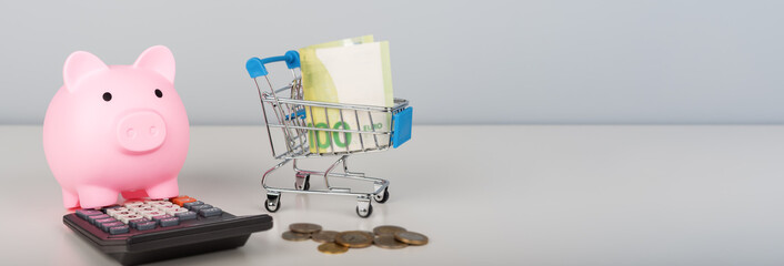 A pink piggy bank, calculator, and miniature shopping cart filled with money.