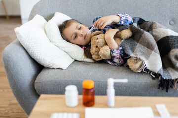 A young girl lies on a couch, taking her temperature with a thermometer, while holding a teddy bear and covered by a blanket. Medications are on the table.