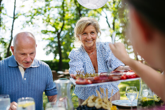 Senior woman serving grilled food during family grill party. - Powered by Adobe