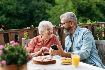 Mature son spending time with elderly mother, showing her something on smartphone.