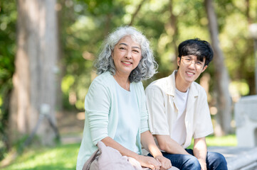 Obraz premium Old asian woman grandmother smiling while sitting on bench with a young man grandson in the park.