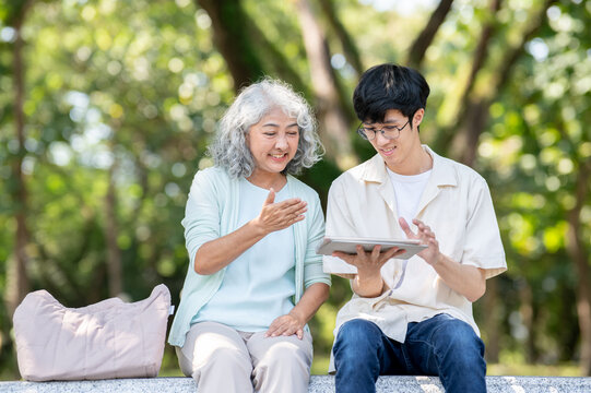 Old asian woman grandmother looking at a young man grandson's tablet while sitting on bench in park.